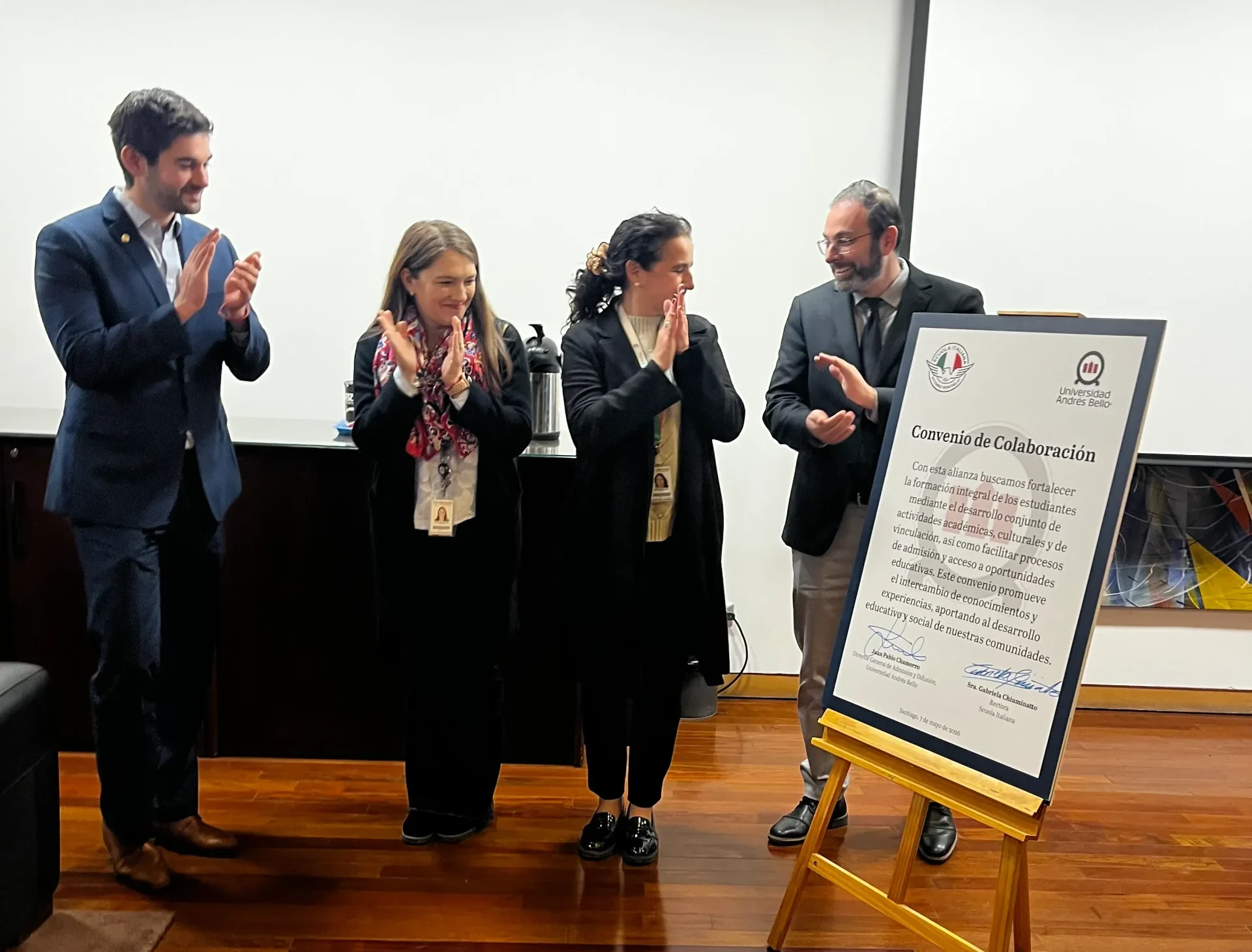 Four professionals in formal attire clapping while standing beside a large board titled 'Convenio de Colaboración'.
