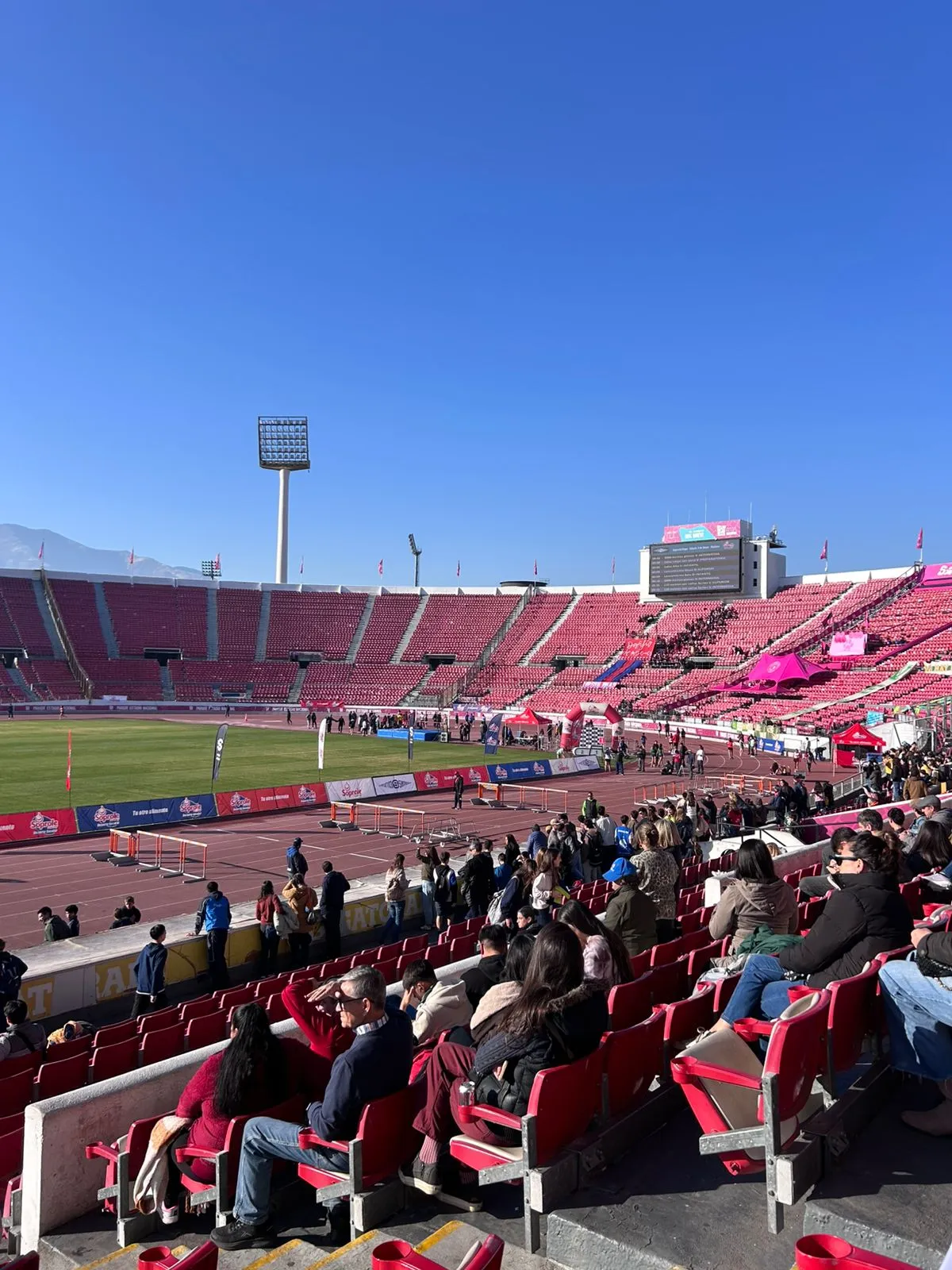 Stadium with red seats and a running track, spectators sit in the foreground on a sunny day with a large scoreboard and tents on the field edge.