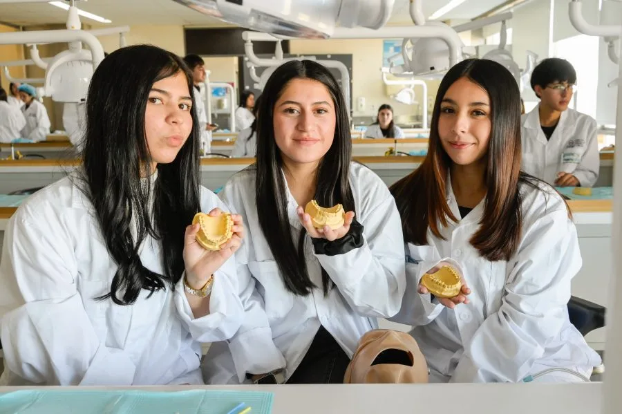 Three young women in lab coats pose in a dental classroom, each holding a dental model in front of a mannequin head on the table.