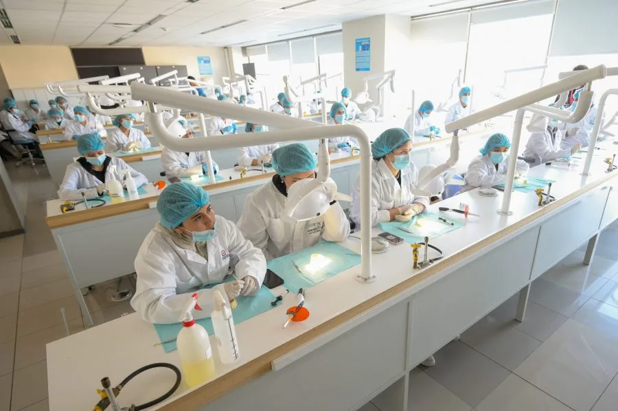 Large laboratory classroom filled with dozens of students in white coats, hair nets, and masks at individual workstations performing lab procedures under bright overhead lights.