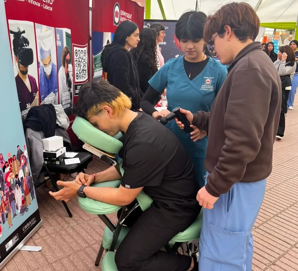 Man in black shirt sits in a green massage chair, resting his chin on a support while a healthcare worker in teal scrubs administers a neck massage at a university fair booth nearby people watch.