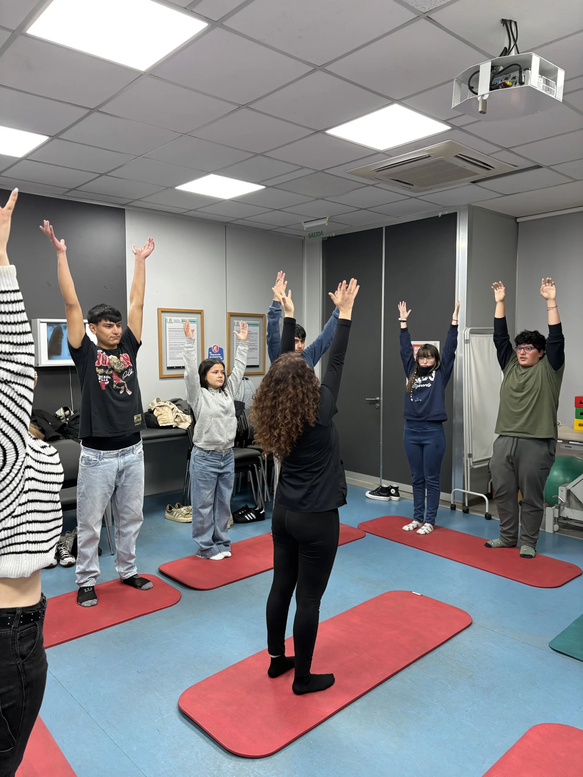 Group of students standing on red yoga mats in a classroom, arms raised during a stretch activity.