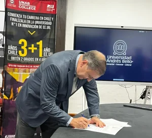 Man in a blazer signs a document on a table during a formal agreement event, with university banners in the background.