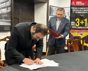 Two men in a conference room: one signs a document on a table while the other stands with a red folder, banner in background indicating a college event.