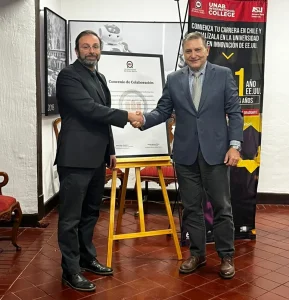 Two men in suits shake hands in a formal signing moment, with a framed document and a red banner behind them.
