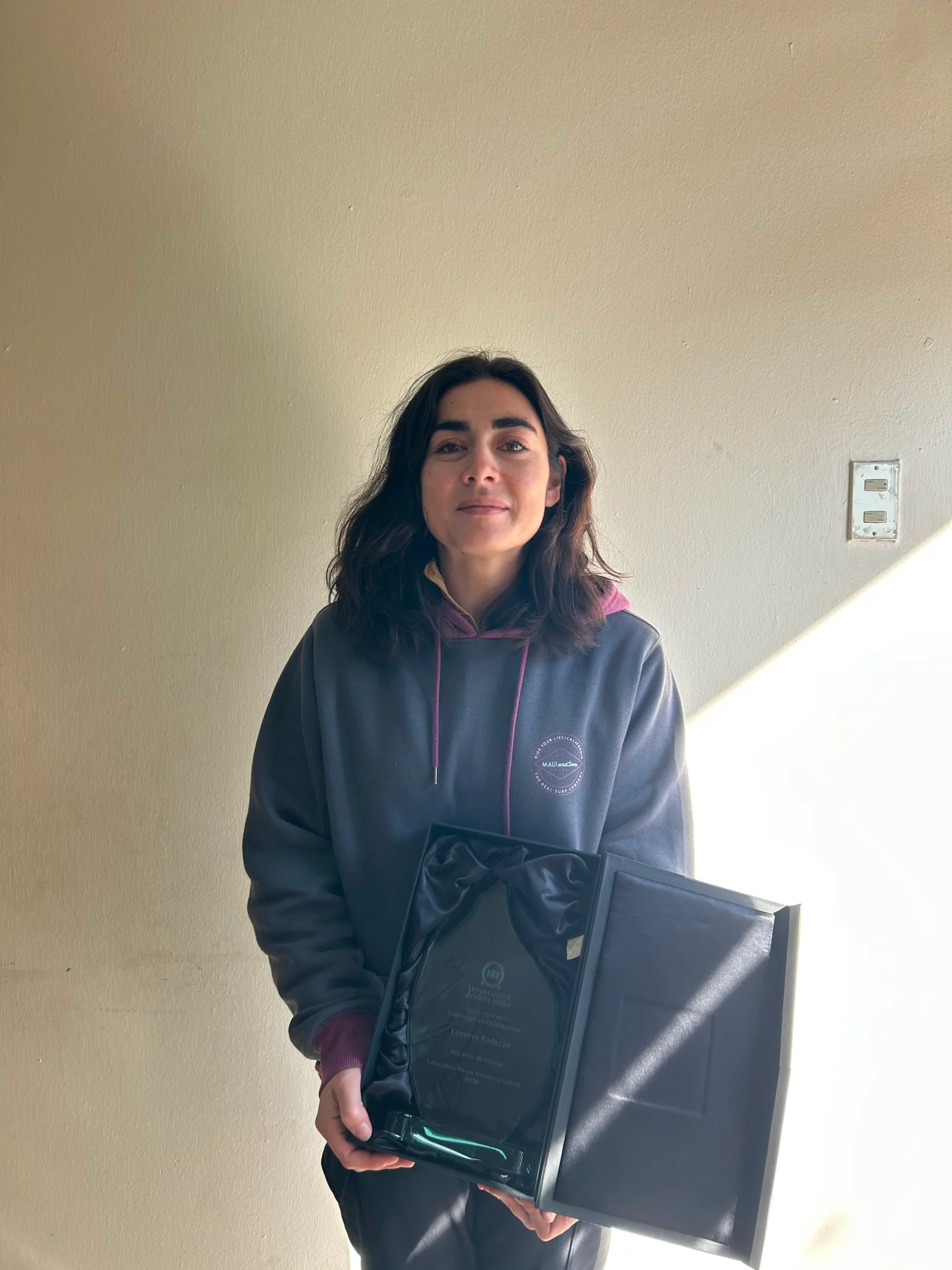 Woman standing indoors against a beige wall, holding a glass award and a black folder, wearing a dark hoodie with pink lining.