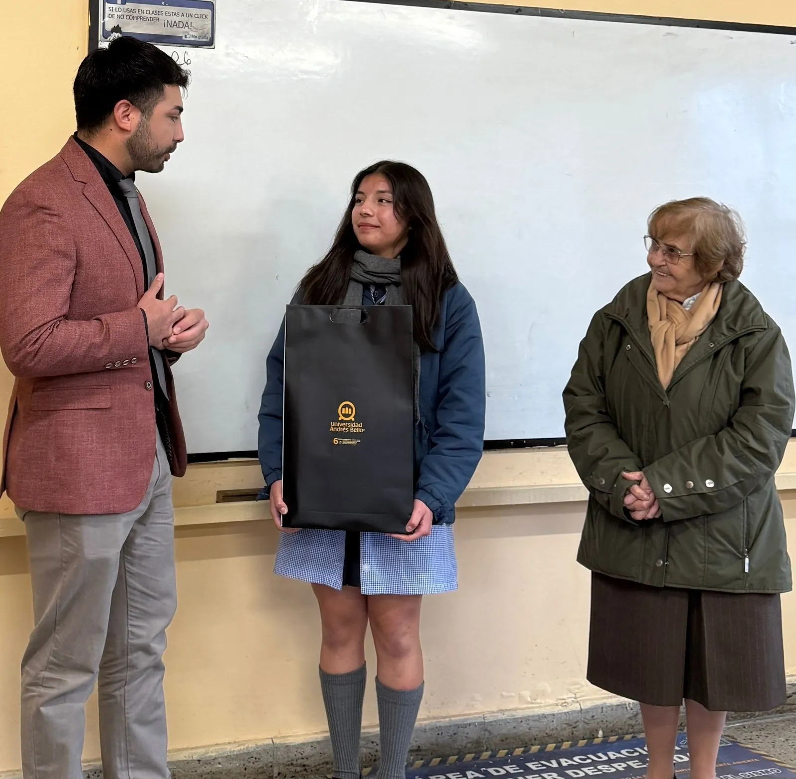 Three people stand in a classroom: a man in a pink blazer speaks with a girl holding a black gift bag, as an older woman smiles nearby.