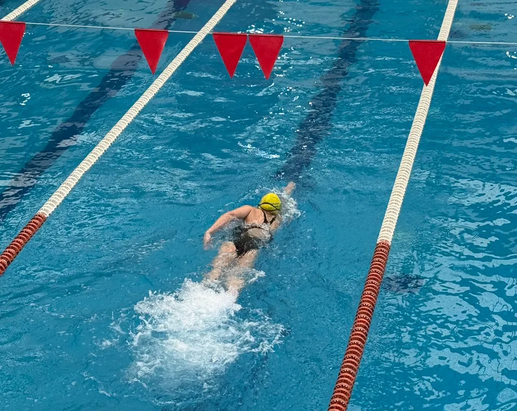 Swimmer in a black swimsuit and yellow cap swimming freestyle in a blue pool lane beneath red triangular flags.