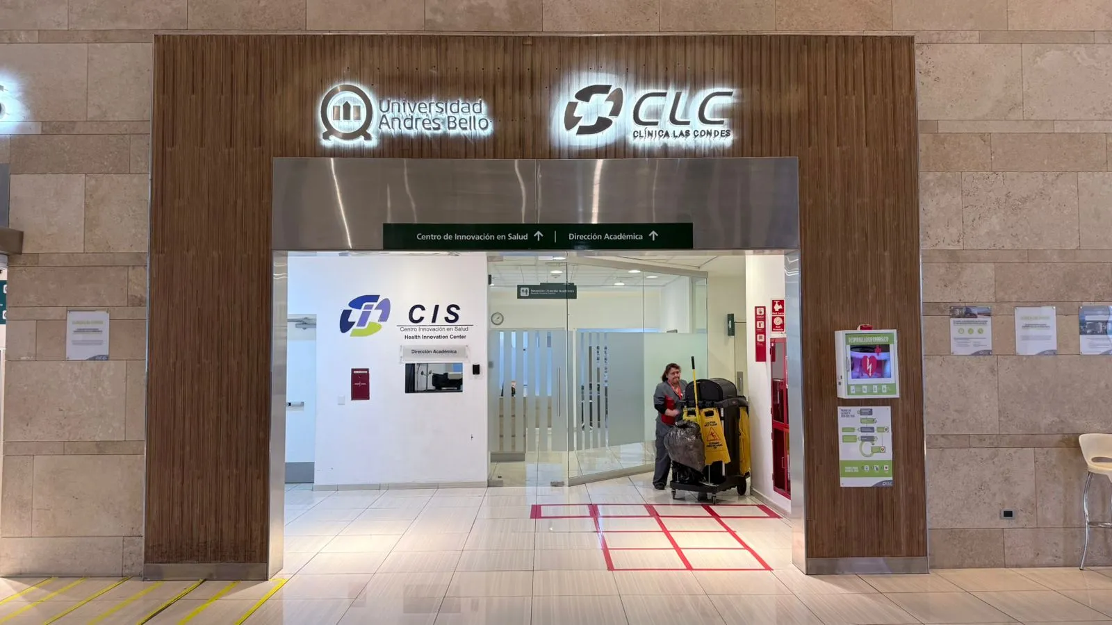 Entrance to the Health Innovation Center (CIS) with glass doors, university logos, and a janitor pushing cleaning equipment nearby