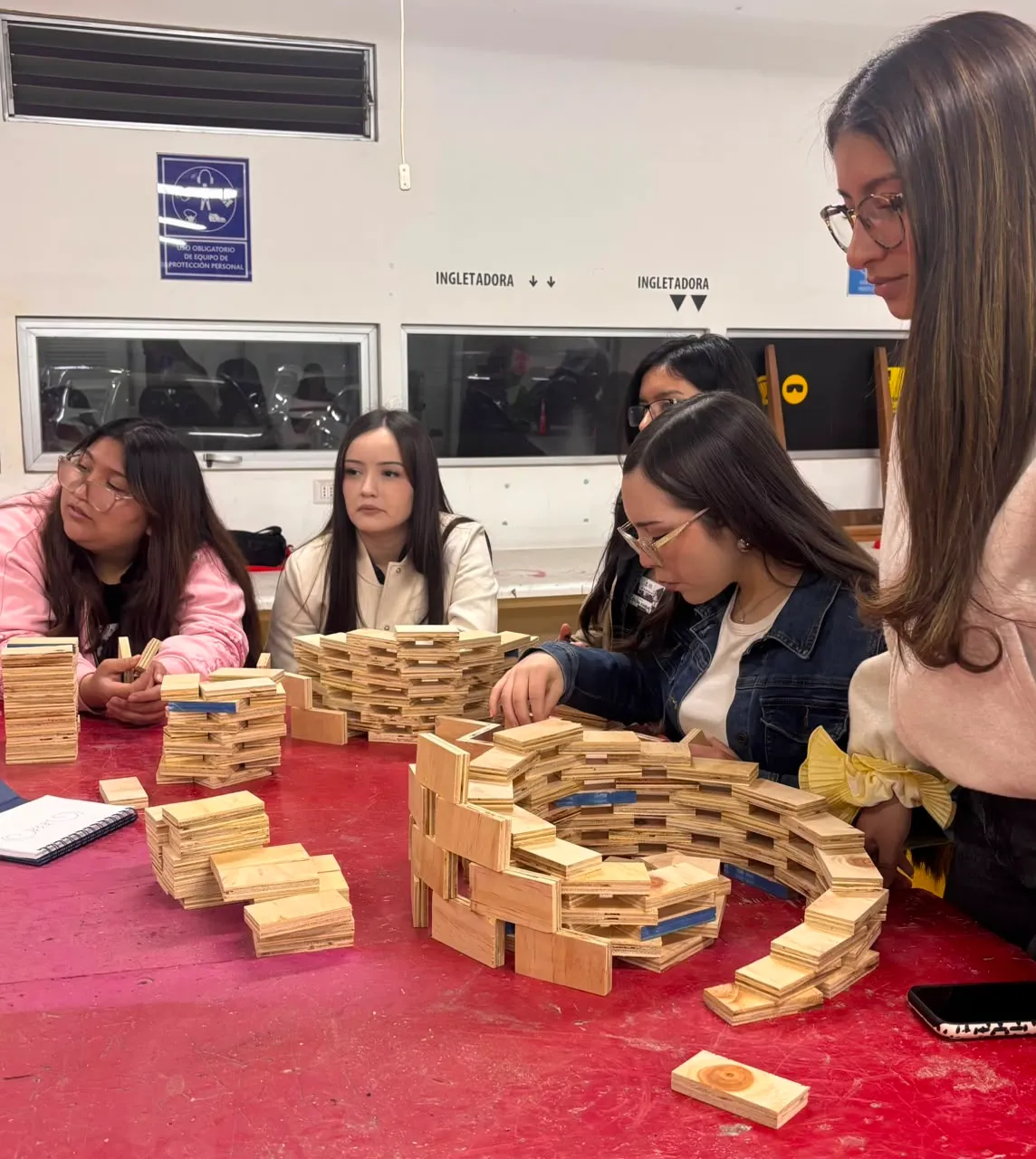 A group of students sits around a red table, stacking small wooden blocks to build multi-layered spiral structures.