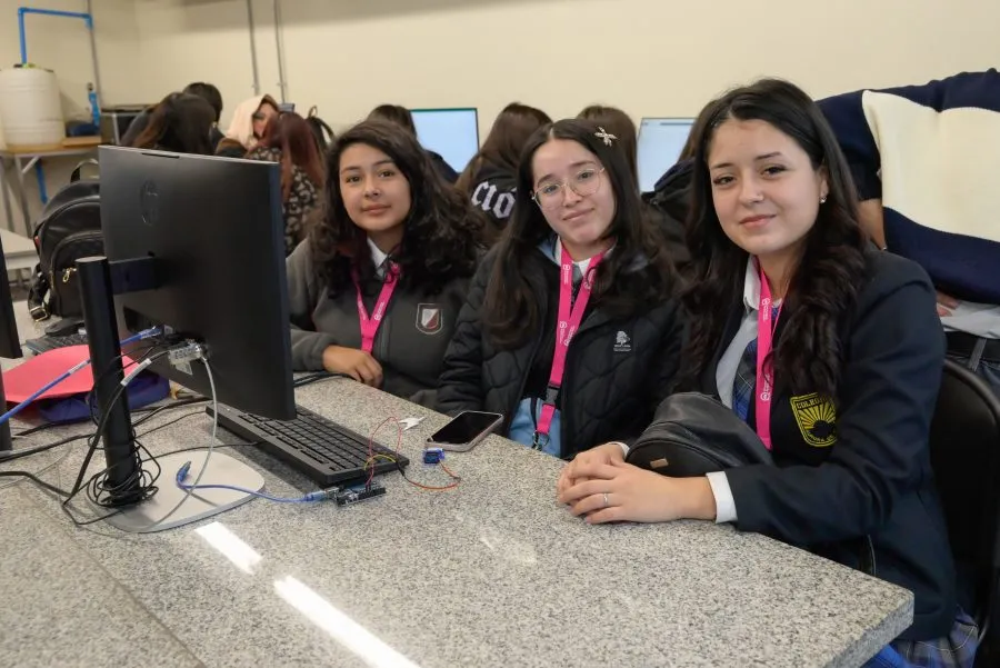 Three students sit at a lab desk in front of a computer, wearing pink lanyards and school uniforms, posing for the camera.