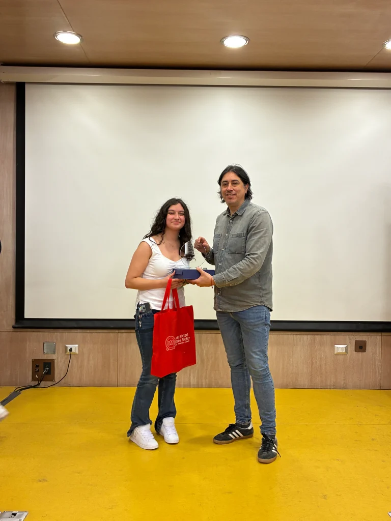 Two people stand on a stage in front of a white screen, exchanging a small box and a red tote bag; both smile.
